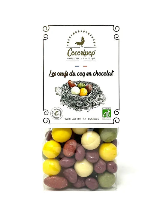 Oeufs du coq en chocolat, d’œufs mignons en chocolat, mélange de graines enrobées aux couleurs variées : noisette chocolat noir verte, soufflé chocolat blanc et mangue jaune, raisin chocolat noir violet et amande fraise rose, présentés sur fond blanc. BIO